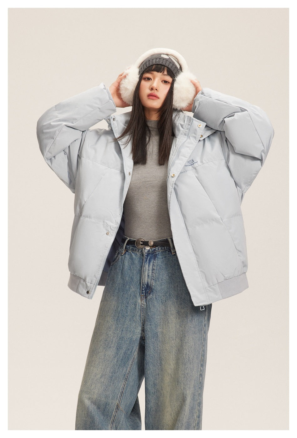 Model wearing a light gray oversized puffer jacket with a high collar, paired with jeans and a knit hat with earmuffs.