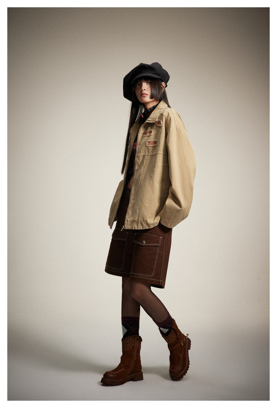 Model in a khaki zip jacket with chest patches, hands in pockets, with brown shorts, black cap and brown boots, side view
