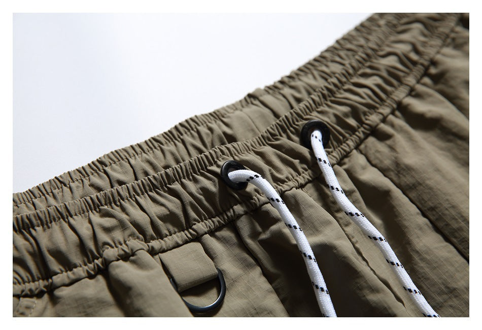 Angled close-up of the gathered waistband, white drawstrings, and black D-ring tab on olive-khaki shorts.