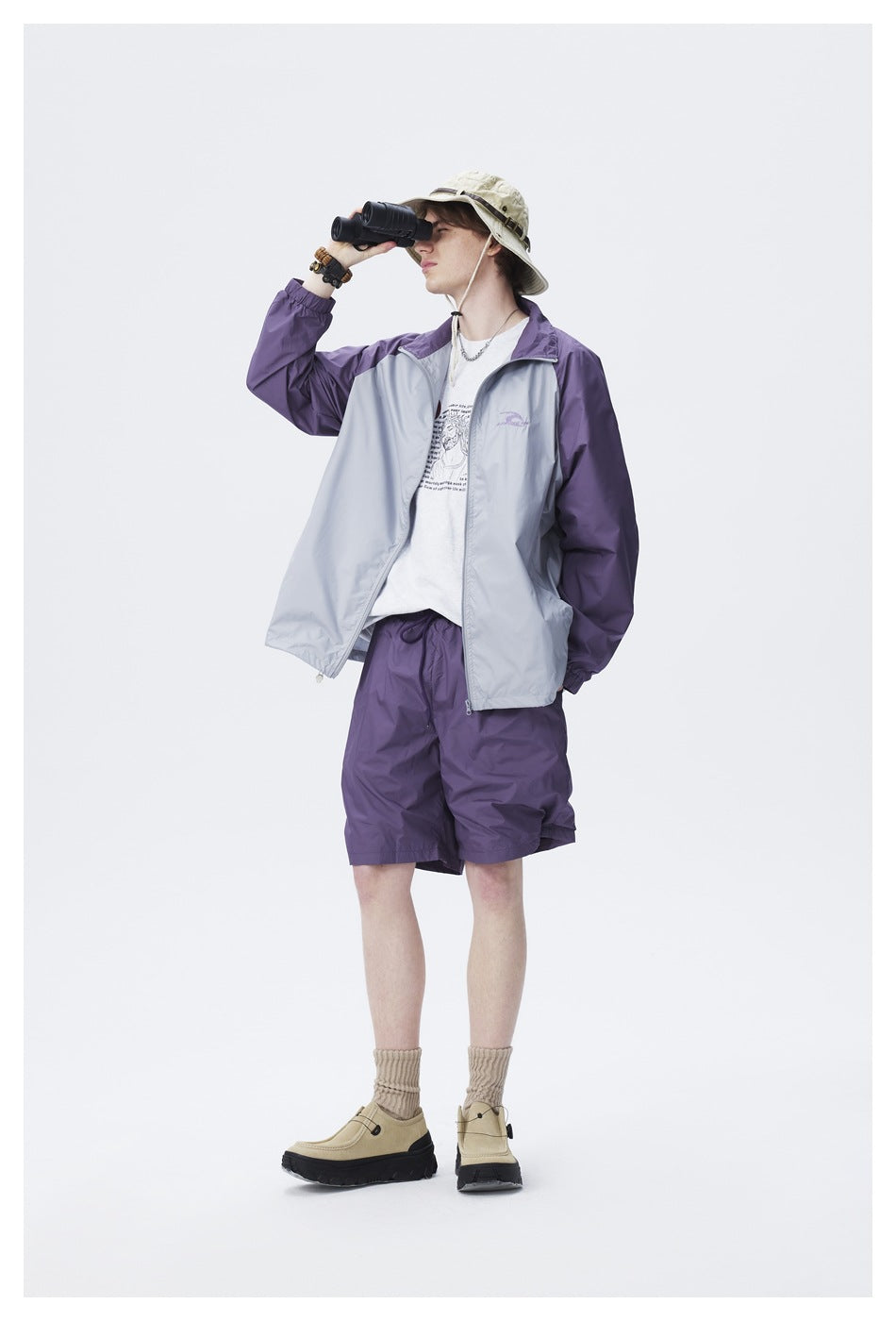 Model wearing an open purple and gray jacket with purple shorts, holding binoculars up to the face