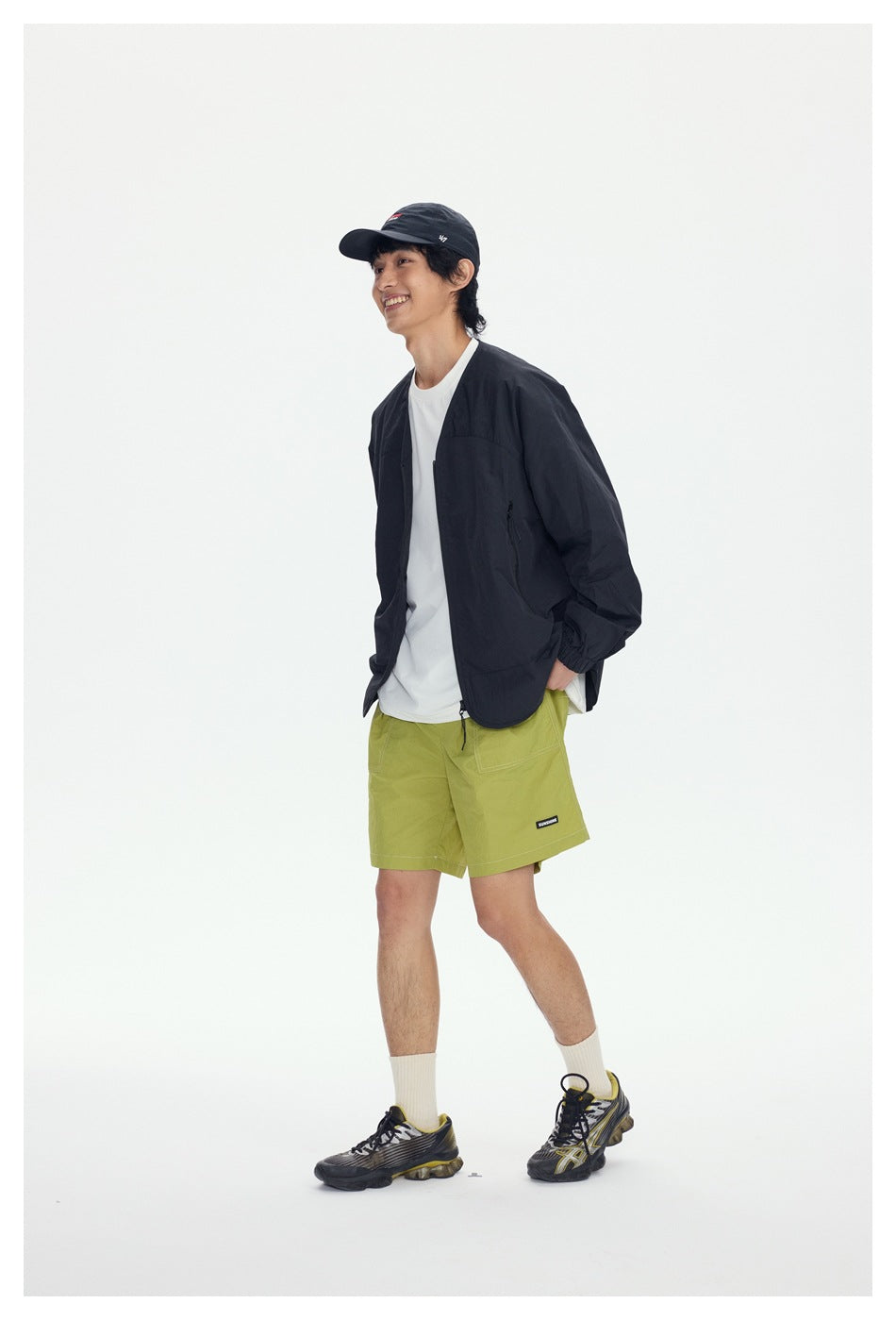 Model in a black V-neck zip jacket worn open over a white tee and green shorts, smiling in a side-angled walking pose
