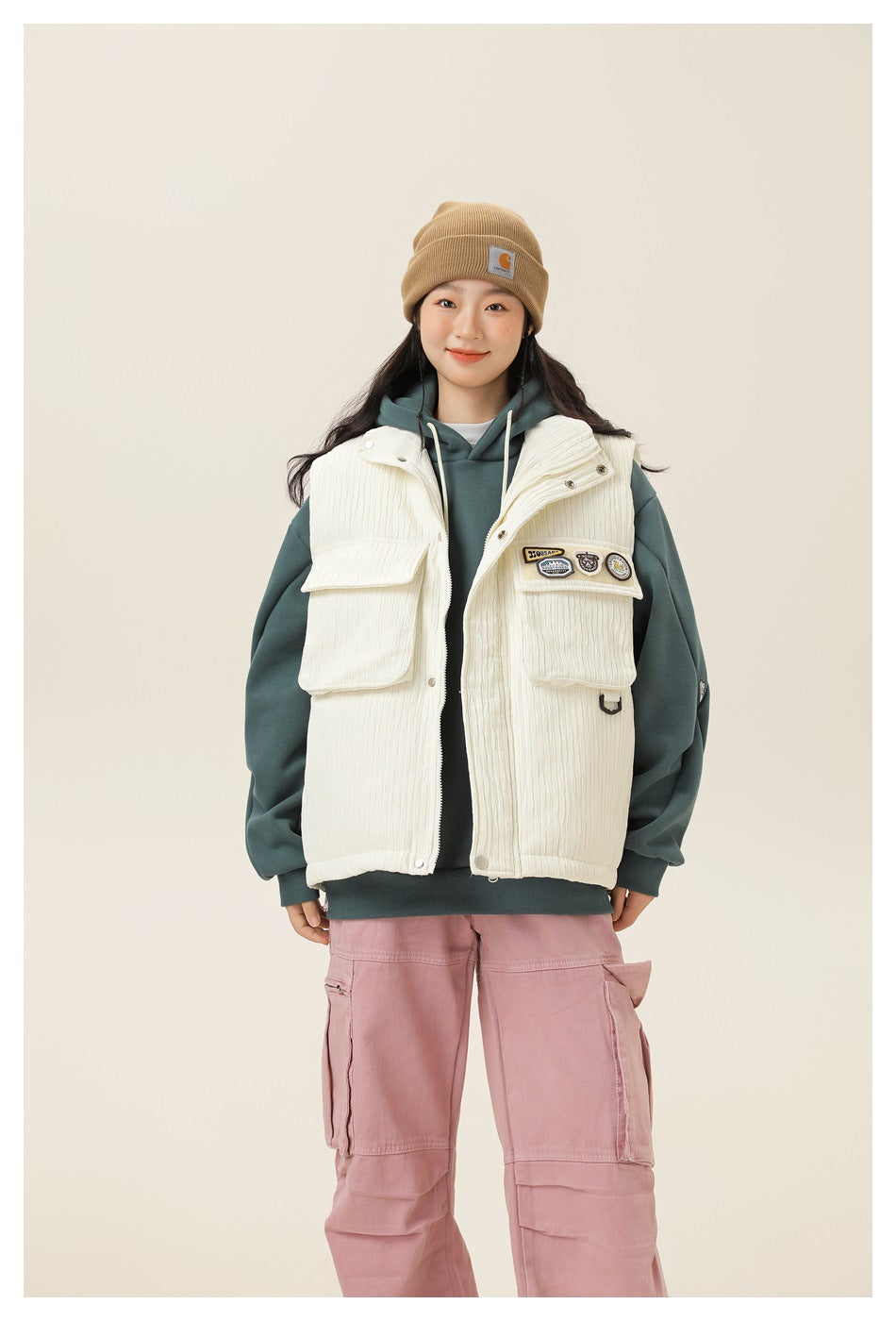 Model wearing off-white textured vest with flap pockets and patches over a teal hoodie, waist-up view