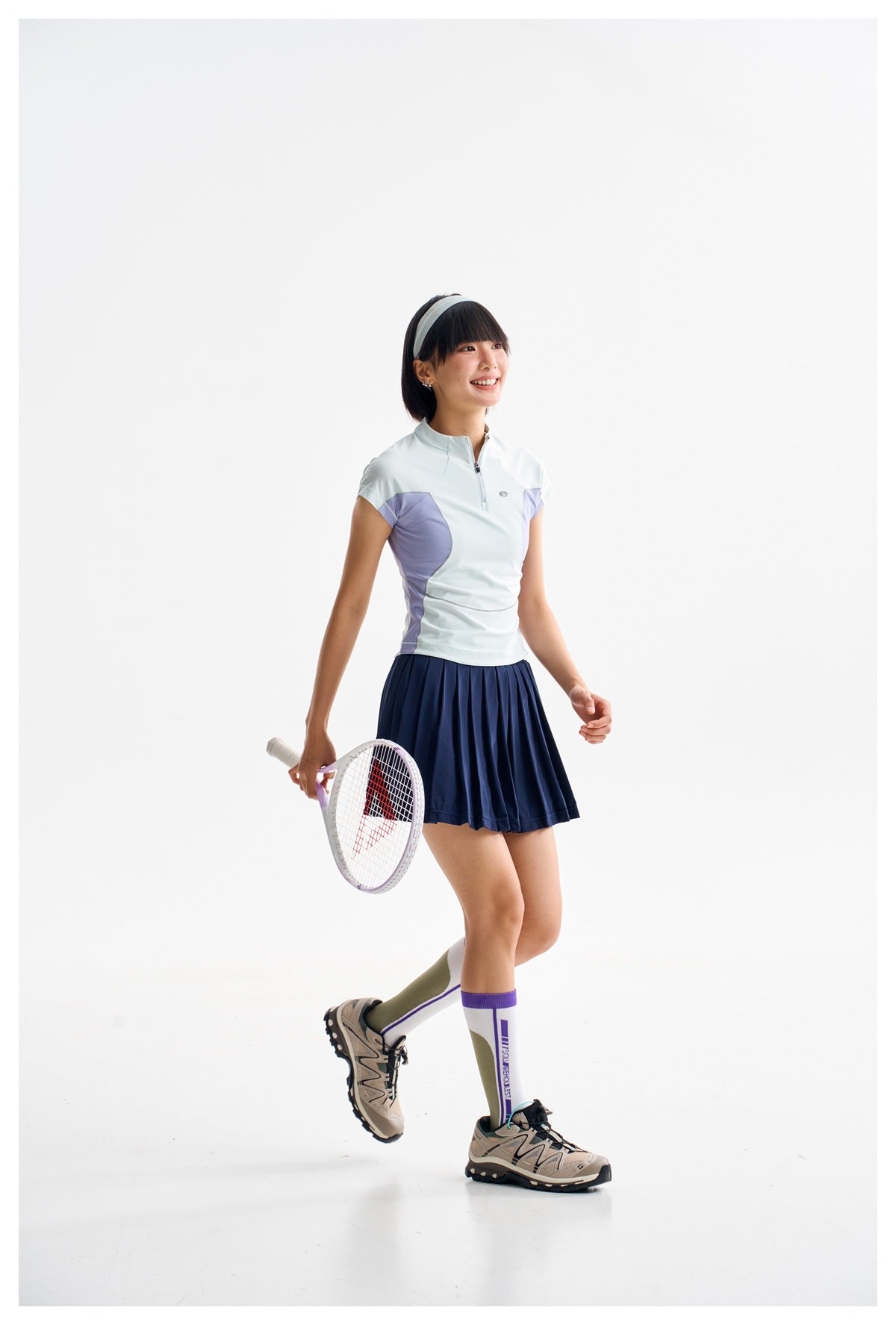 Woman walking with a tennis racket, wearing a pale blue quarter-zip top with lavender panels and a navy pleated skirt.