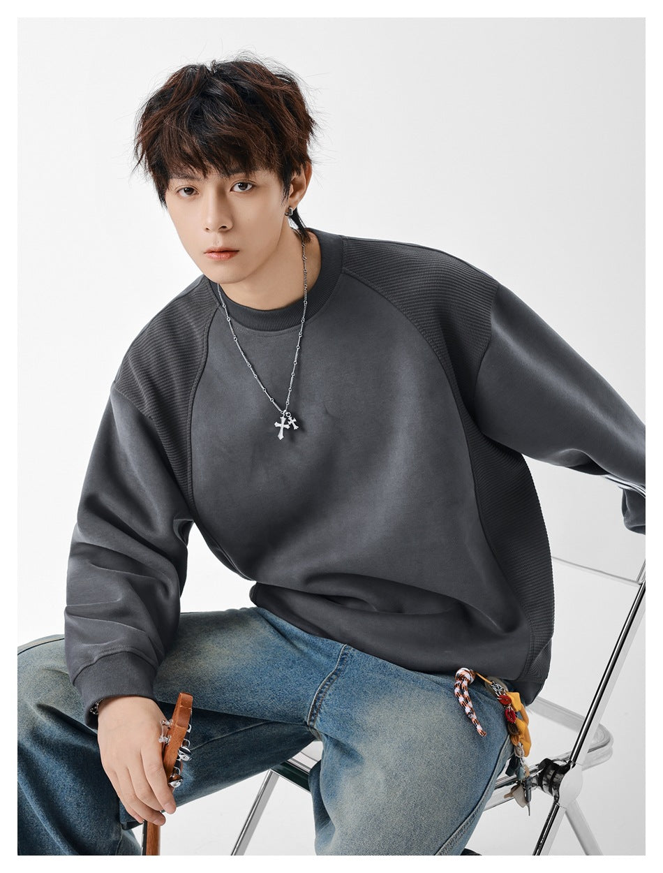 Man seated wearing gray raglan crewneck sweatshirt with ribbed shoulder panels, jeans and a cross necklace