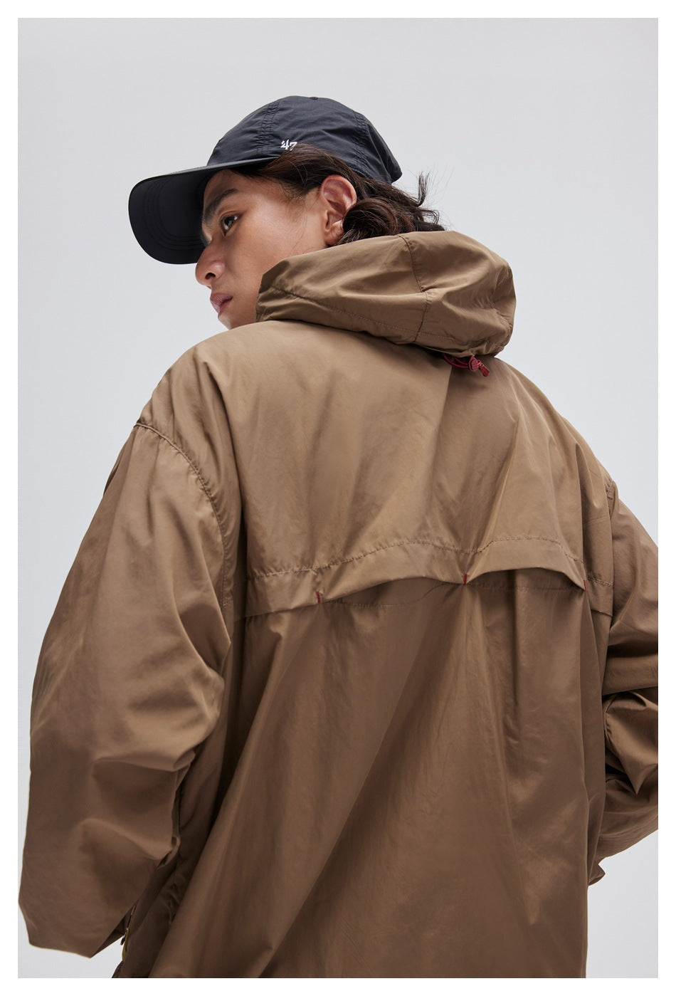 Model looking over her shoulder in a taupe hooded jacket, showing its back seams and hood.