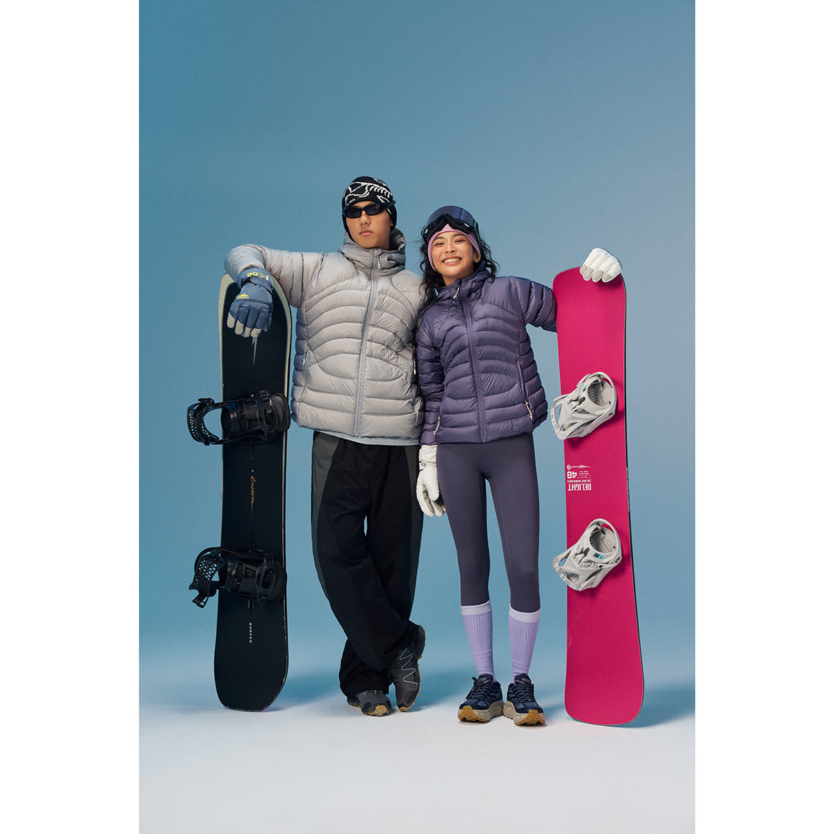 Two models wear light gray and purple puffer jackets beside upright snowboards against a blue backdrop.