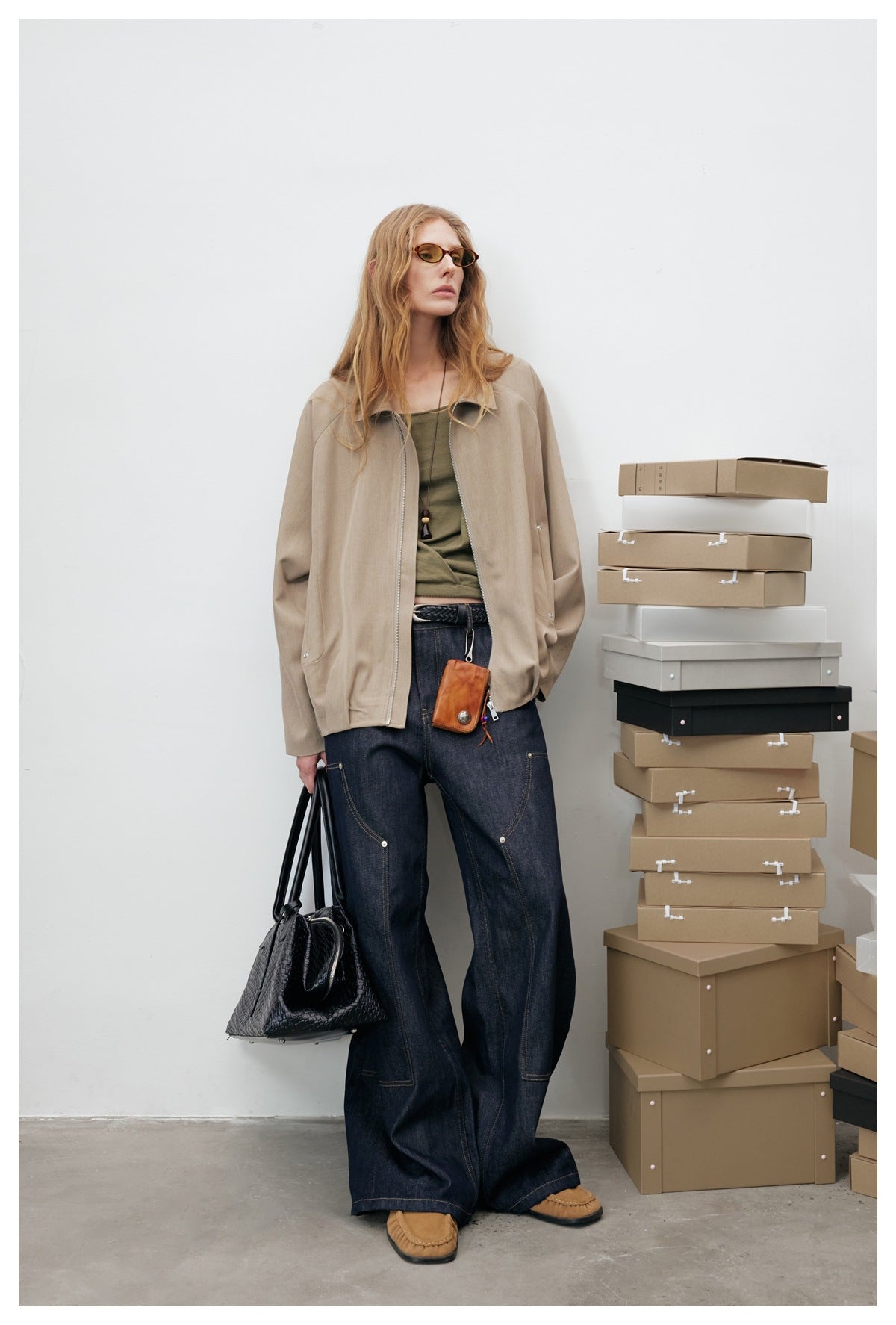 Model leaning on a white wall in an open khaki zip jacket, olive top and wide-leg dark jeans, carrying a black woven bag