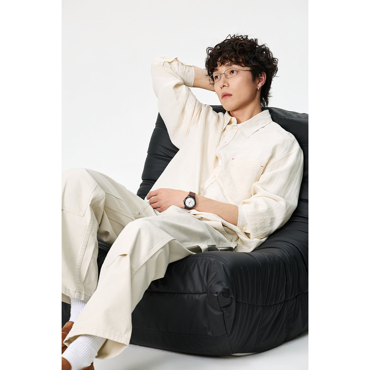 Model seated in a black chair wearing an ivory button-up shirt with a chest pocket and matching relaxed trousers.