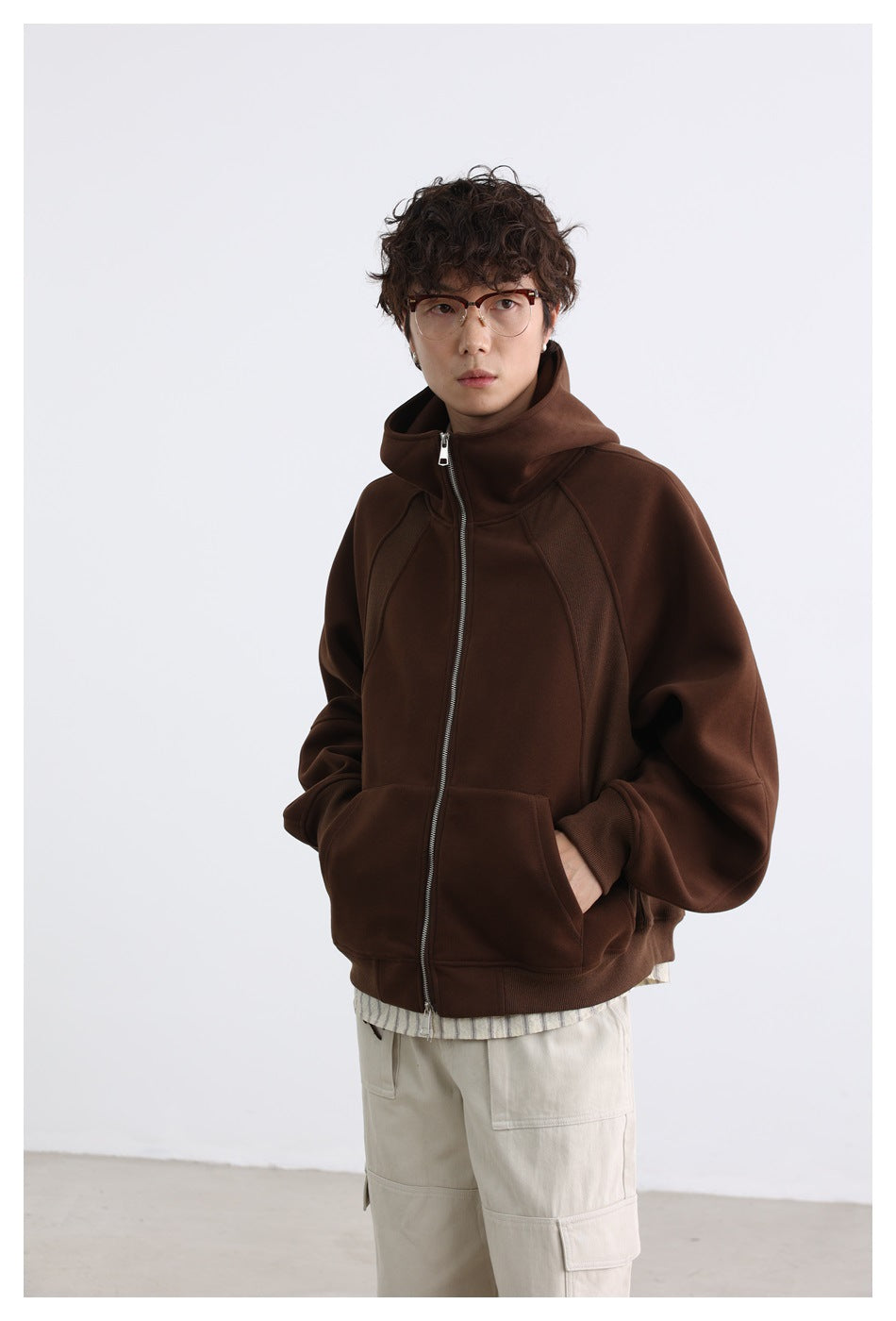 Model wearing a zipped dark brown hooded jacket with a high collar, raglan seams, and front pouch pockets.