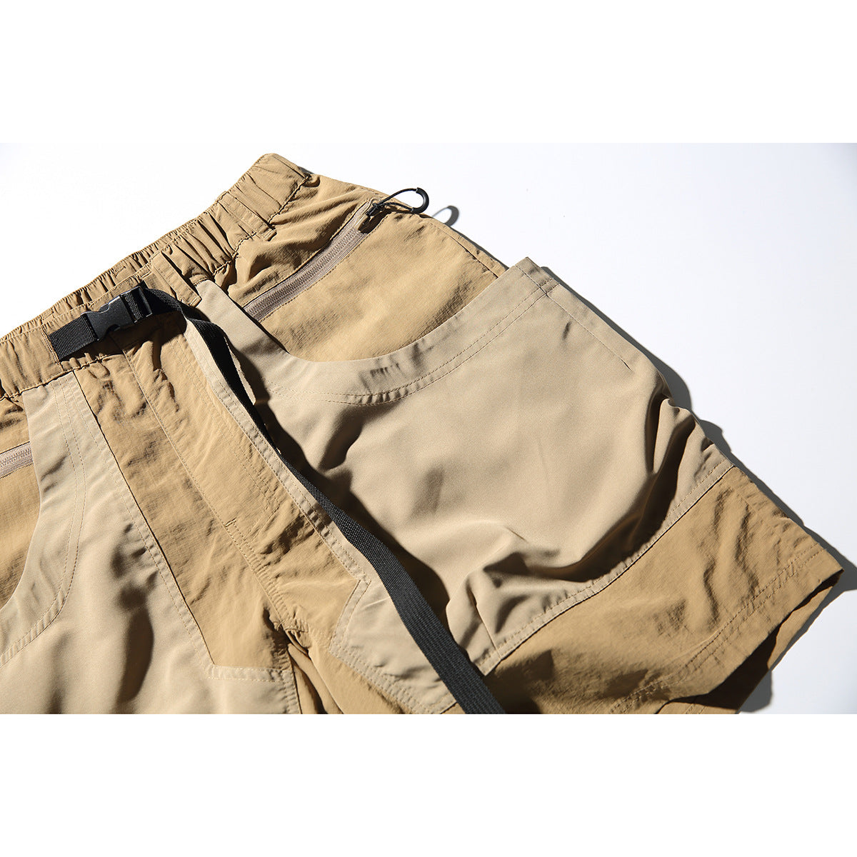 Angled close-up of tan shorts showing a large curved pocket, zippered pocket, and black belt strap.