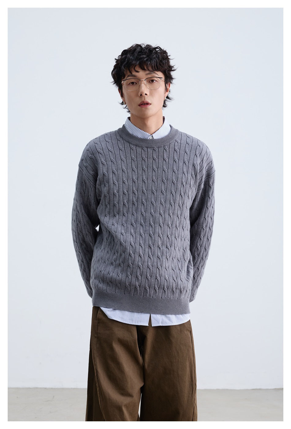 Front view of a model wearing a gray cable-knit crewneck sweater over a striped shirt and brown trousers.