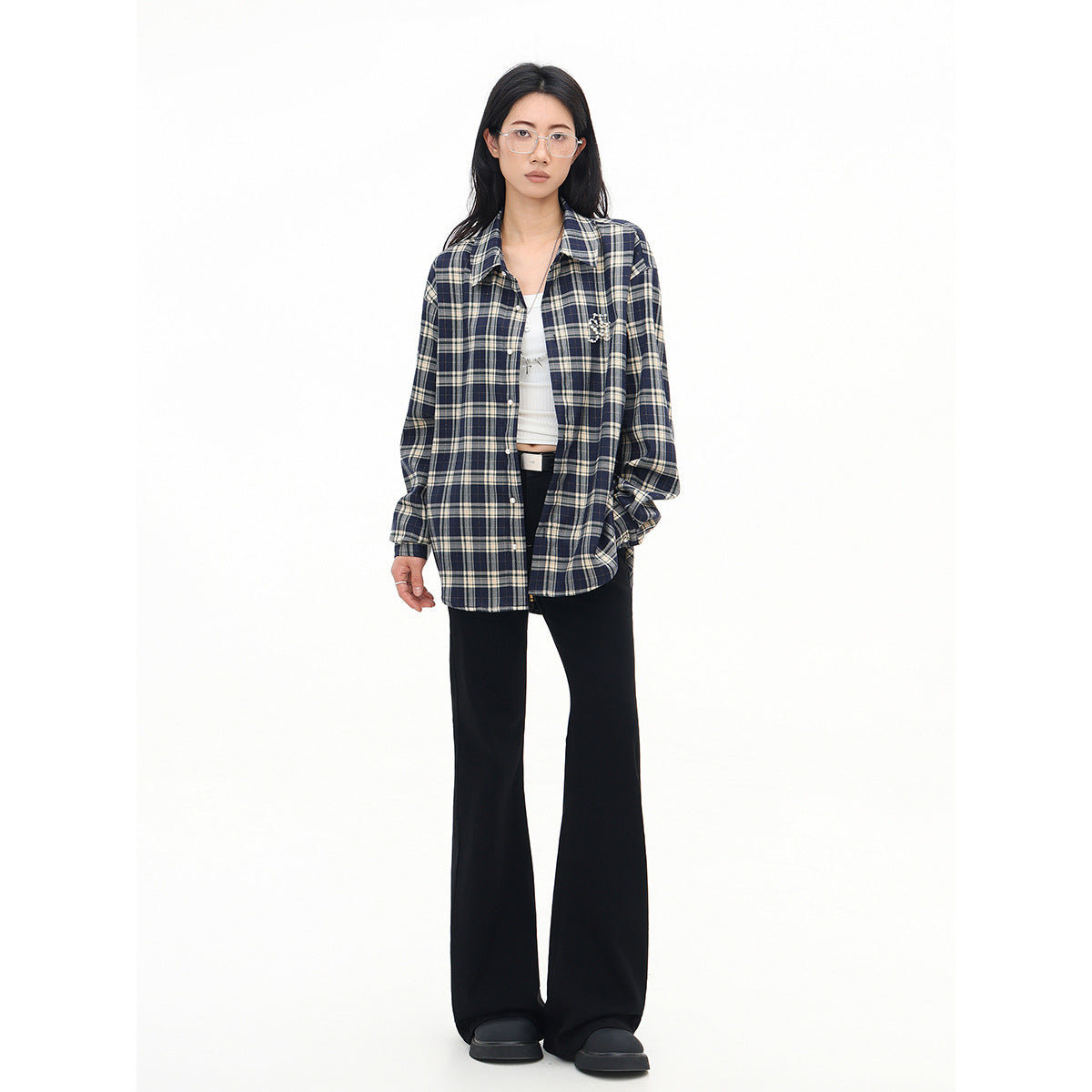 Model wearing an open navy and cream plaid shirt with a beaded chest embellishment, white crop top, and black flared pants.