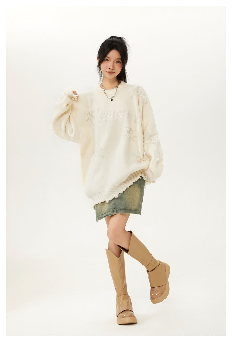 Model posing in an ivory distressed sweater with tonal chest lettering, a faded denim skirt and tan boots.