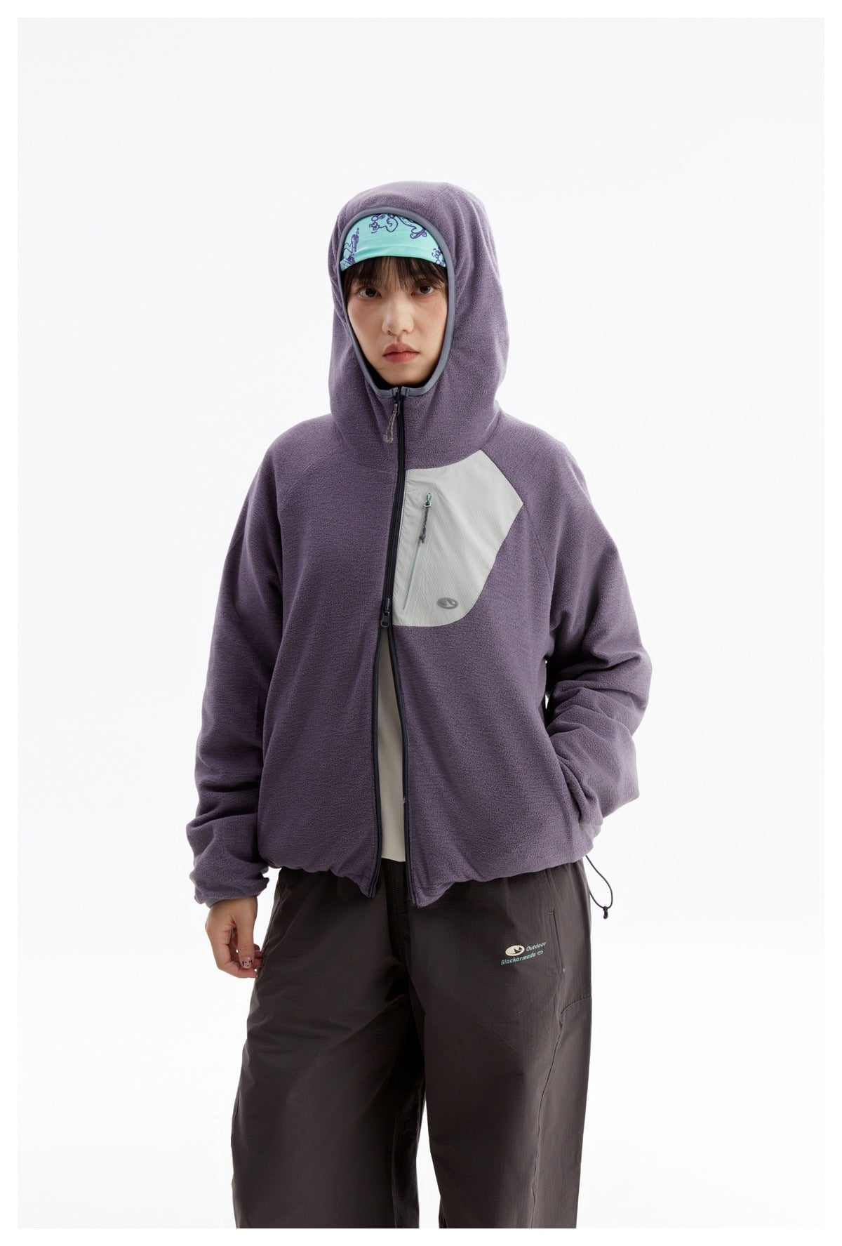 Model wearing a purple jacket with the hood raised, face opening trimmed in gray, and a pale zip chest pocket.