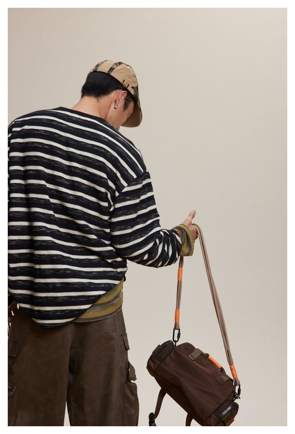 Rear view of a model wearing a loose black-and-cream striped long-sleeve shirt with brown trousers and a tan cap.