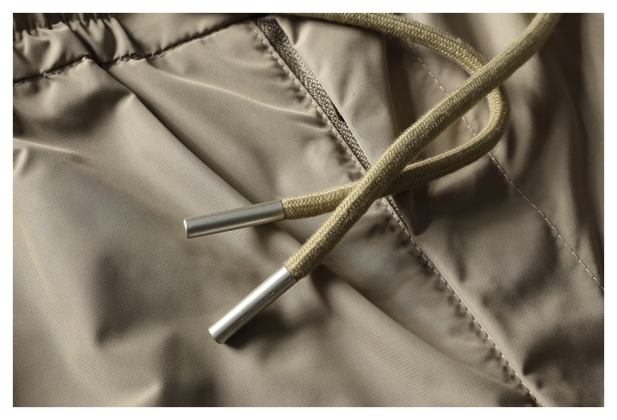 Close-up of taupe trousers with a braided drawstring, metal tips, and a slanted pocket opening.