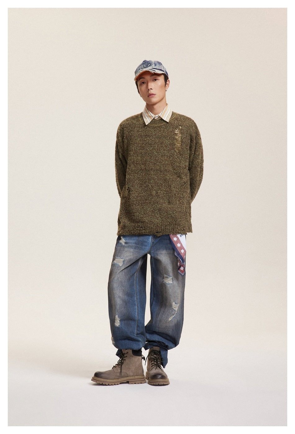 Full-length front view of a model in an olive-brown distressed sweater, striped collar shirt, loose jeans and boots.