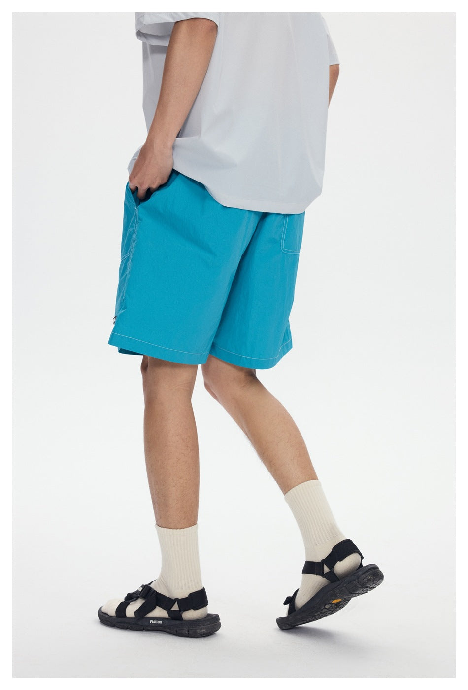 Rear angled view of a model wearing bright turquoise knee-length shorts with stitched pockets and a light gray T-shirt.