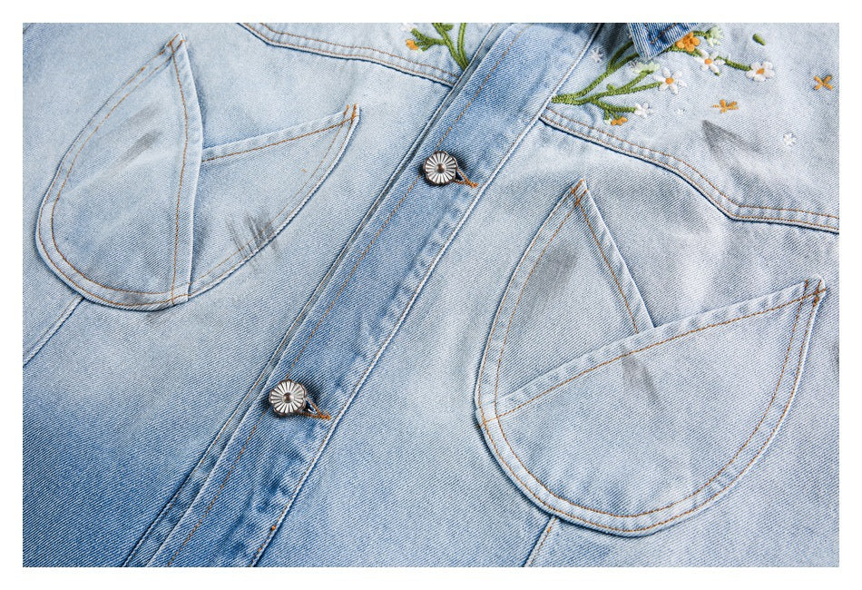 Close-up of a light-wash denim jacket front with curved chest pockets, metal buttons, and floral embroidery.