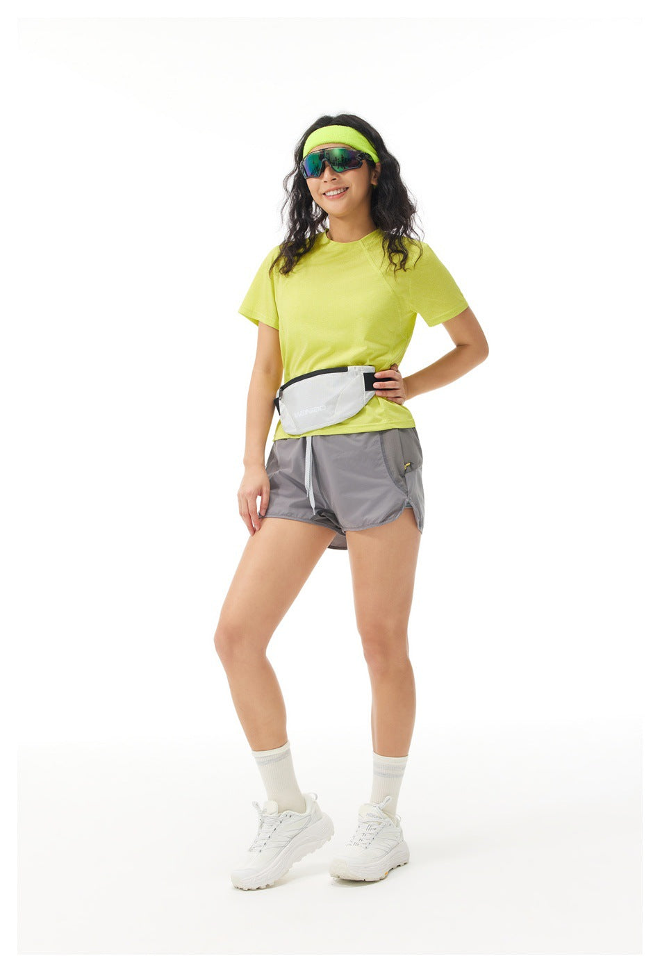 Full-length model wearing a lime shirt, gray shorts, and a light waist pack, standing against a white background.