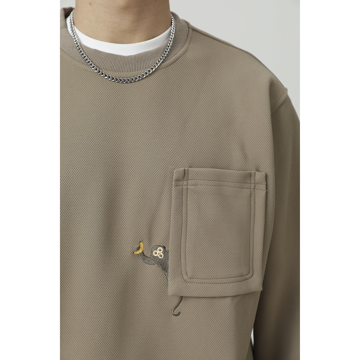 Close-up of the taupe sweatshirt's textured fabric, chest pocket, small embroidered motif, and ribbed neckline.