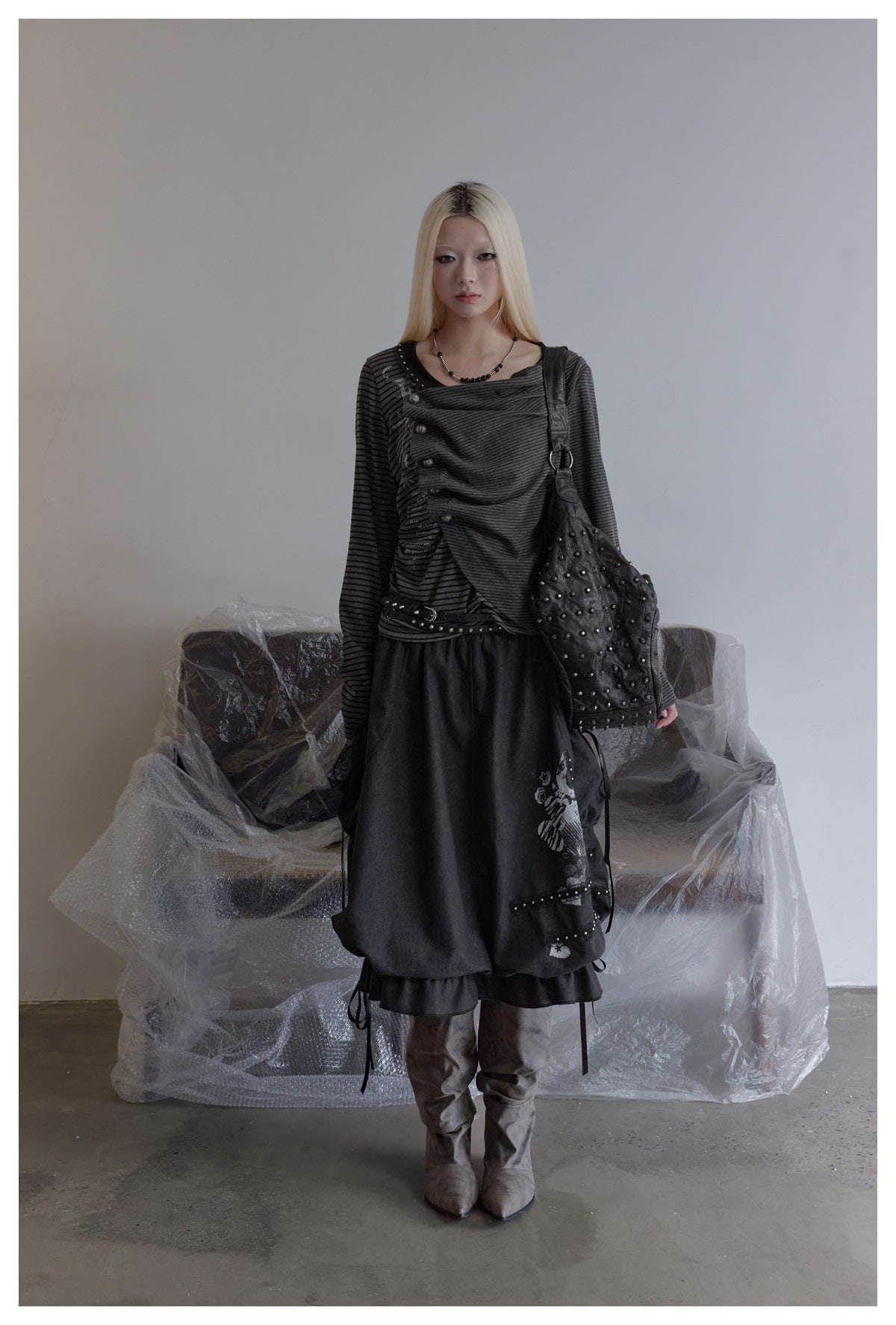 Model wearing the gray striped asymmetrical button-placket top with a dark skirt, studded bag and boots in front of a sofa