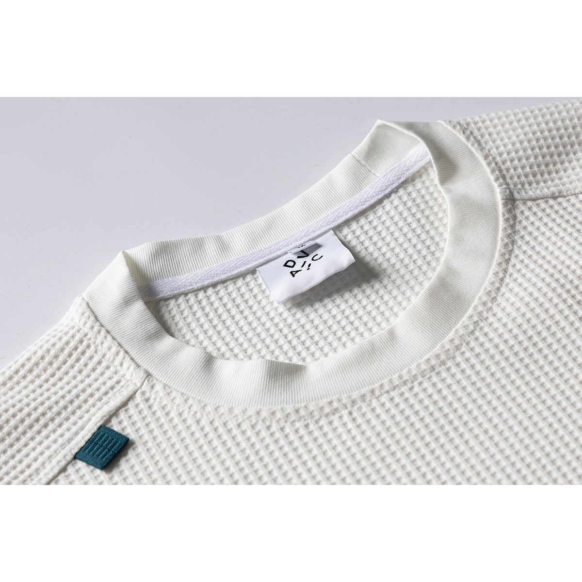 Close-up of an ivory waffle-textured shirt's ribbed crew neck, inner label, and small teal fabric tab.