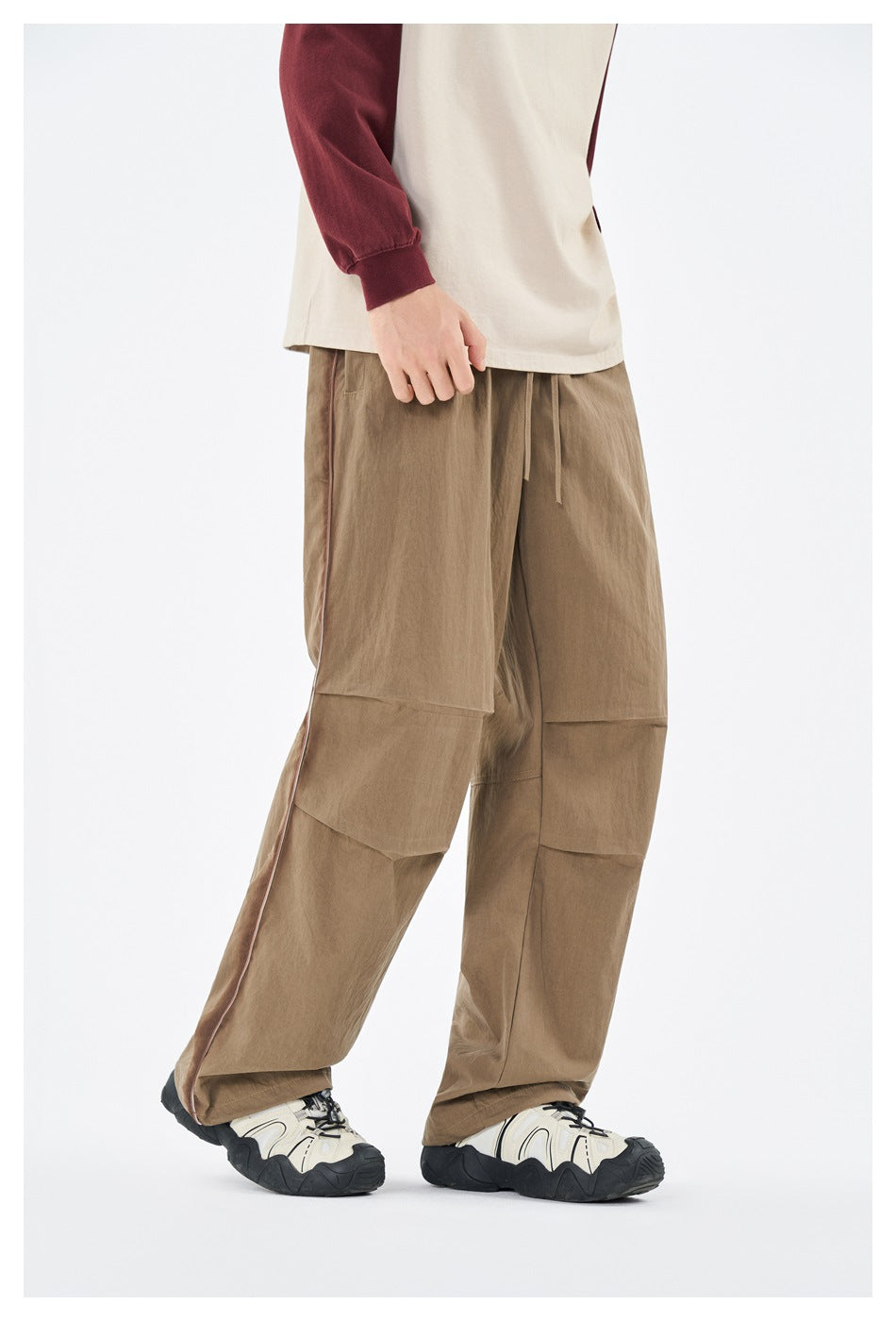 Front angle of a model wearing loose khaki pants with a drawstring waist, paneled legs, and contrast piping.