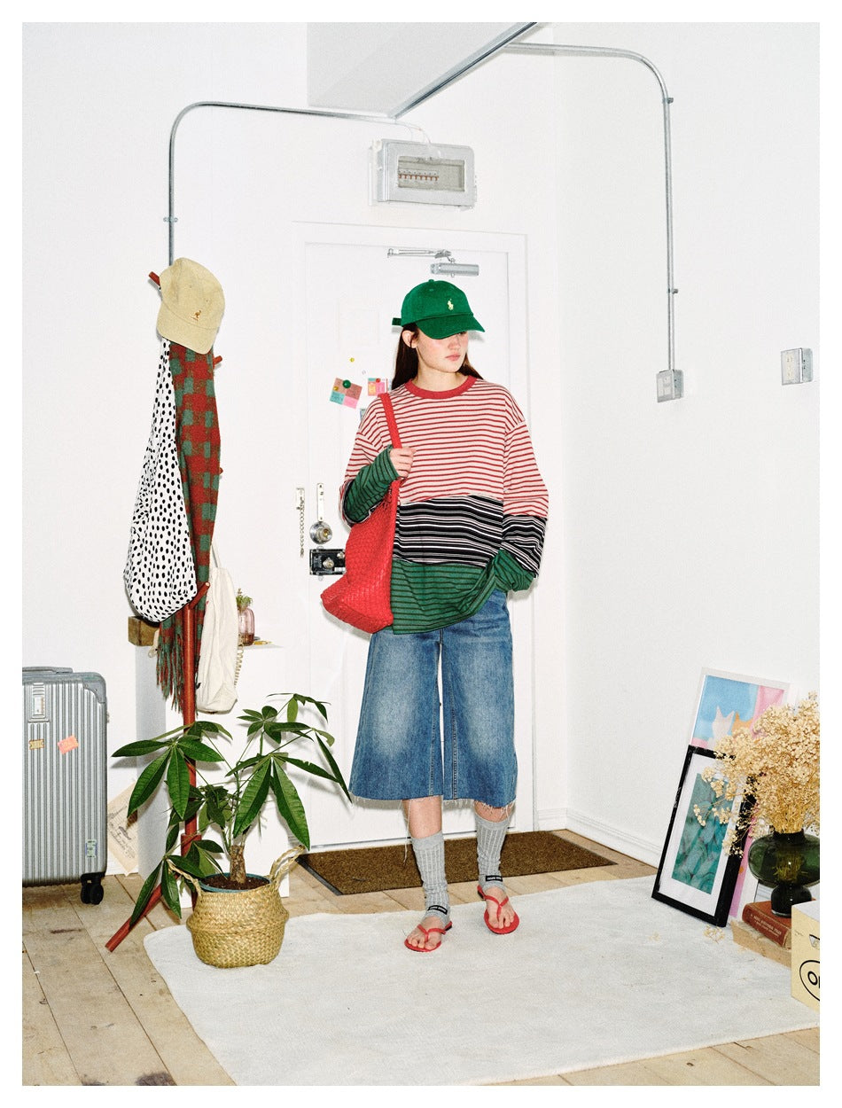Model in a striped patchwork long-sleeve top with denim shorts, green cap and red tote, turned slightly to the side