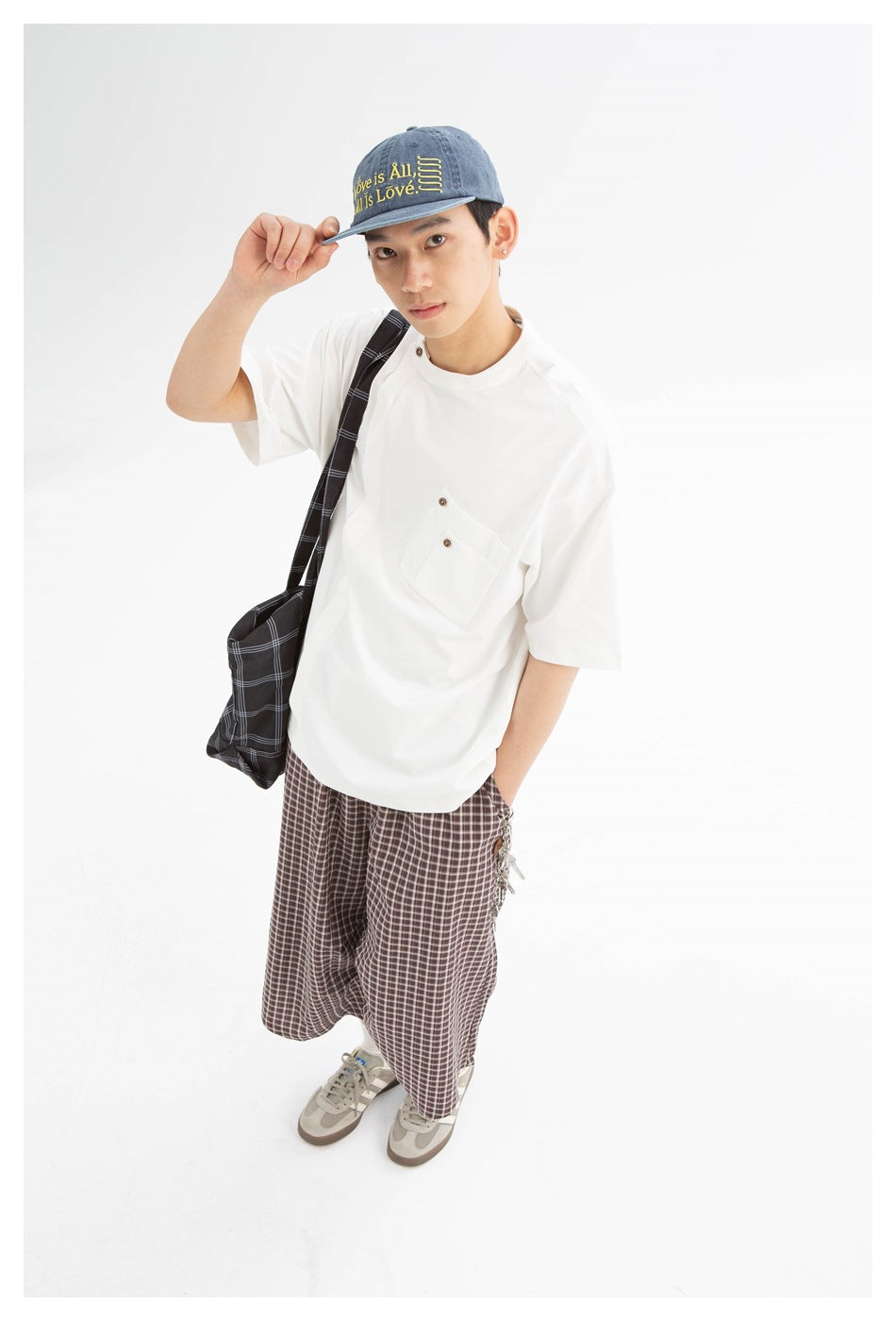 Model wearing an oversized white short-sleeve pocket T-shirt, plaid trousers, and a blue cap with yellow lettering.