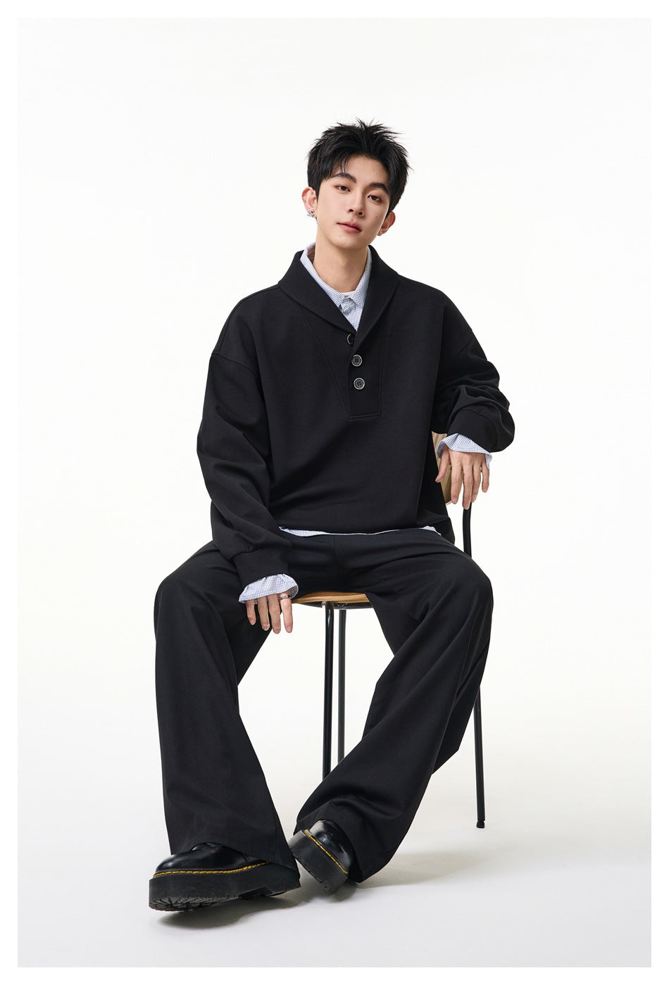 Seated model wearing a black oversized three-button pullover over a checked shirt with wide-leg black trousers.