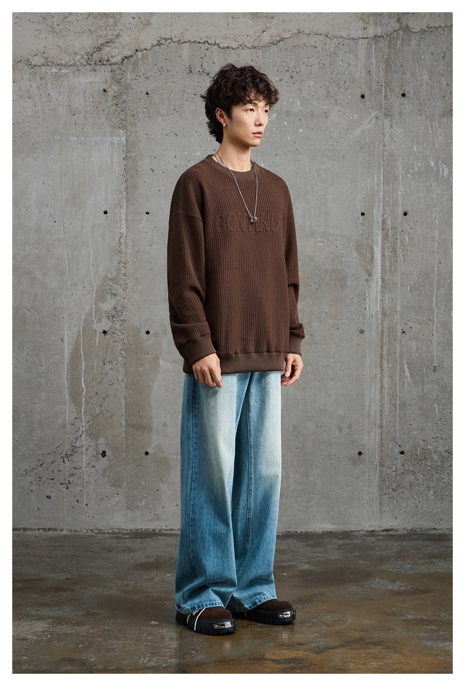 Model wearing an oversized brown textured crewneck sweatshirt with subtle raised chest lettering and light blue wide-leg