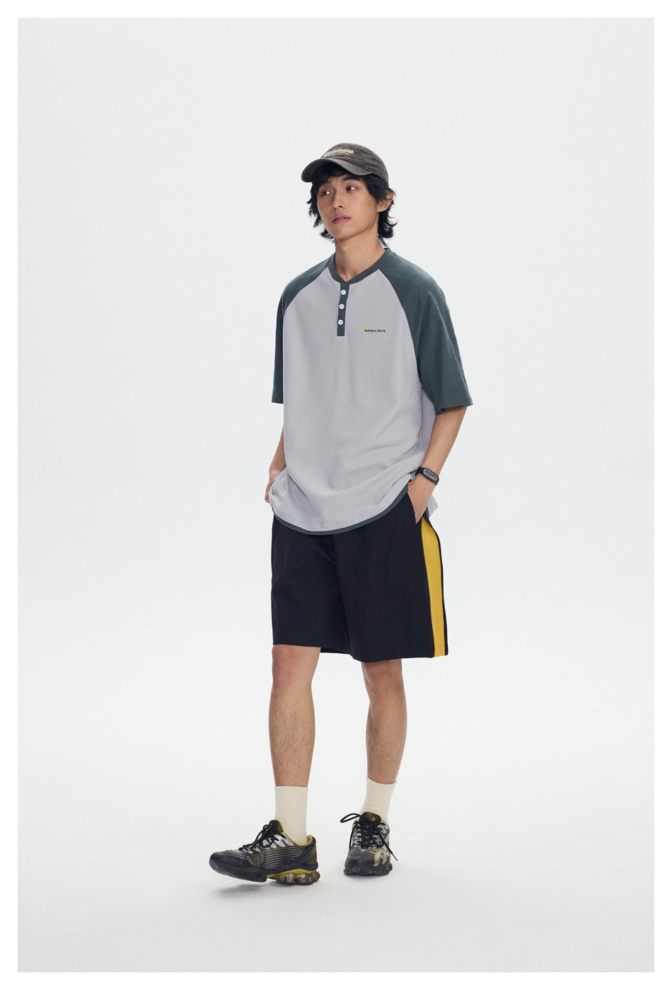 Full-length view of a model wearing a light gray henley with dark green raglan sleeves and black shorts with a yellow side