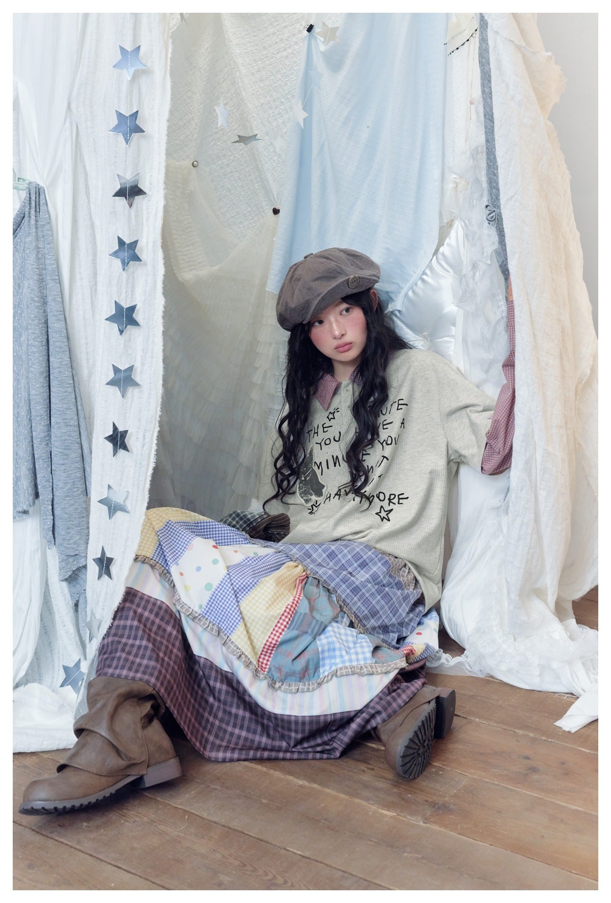 Seated model wearing a cream graphic top with red checked sleeves and a voluminous multicolored patchwork skirt.