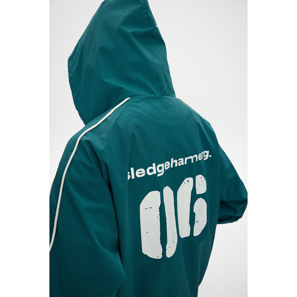 Close rear view of a teal hooded jacket with cream shoulder piping and large white lettering and graphic on the back.