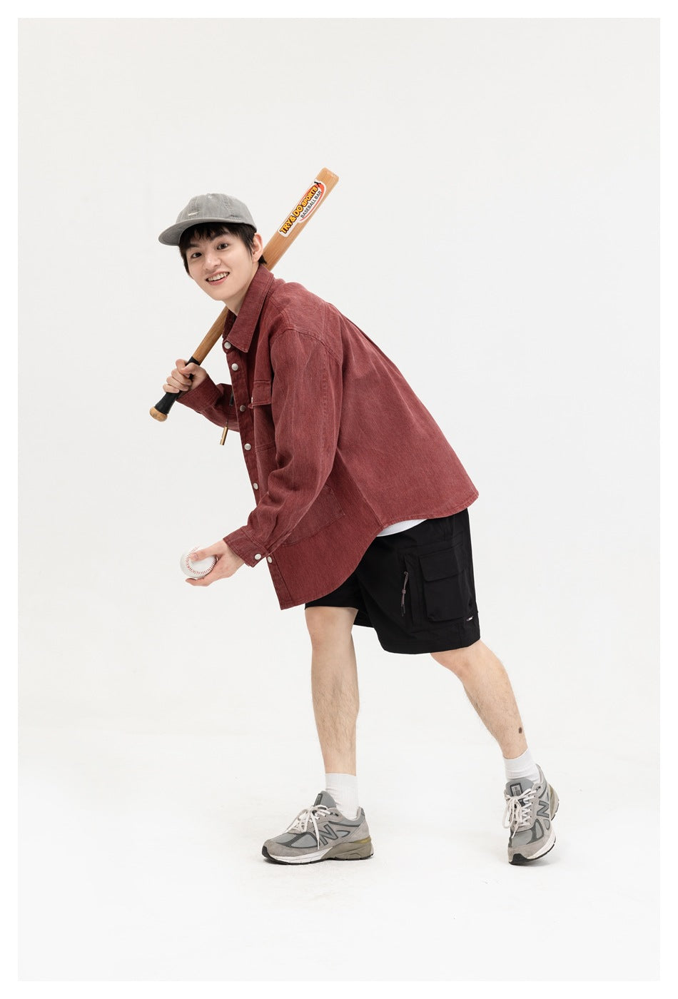 A man wears a burgundy button-front overshirt and black cargo shorts while holding a baseball bat and ball.