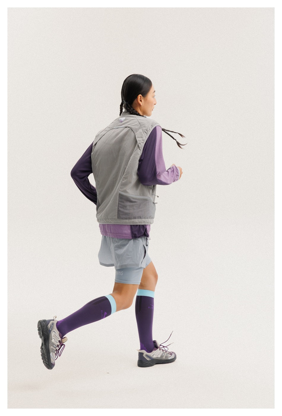 Rear three-quarter view of a model running in a gray vest, purple top, gray shorts, and purple knee-high socks.