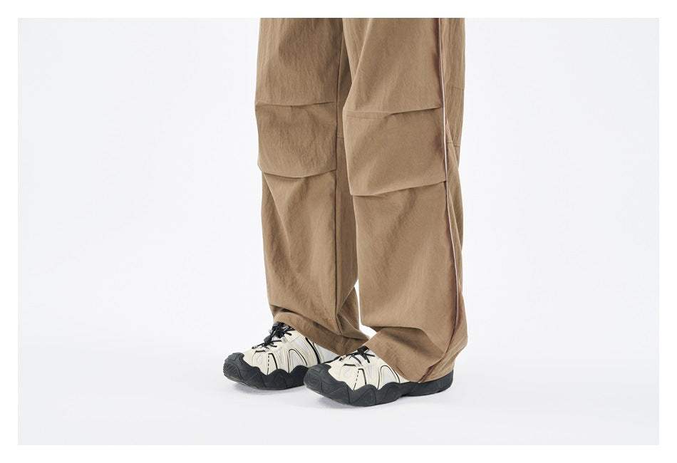 Close view of tan trousers showing paneled knees, wide hems, side piping, and sneakers.