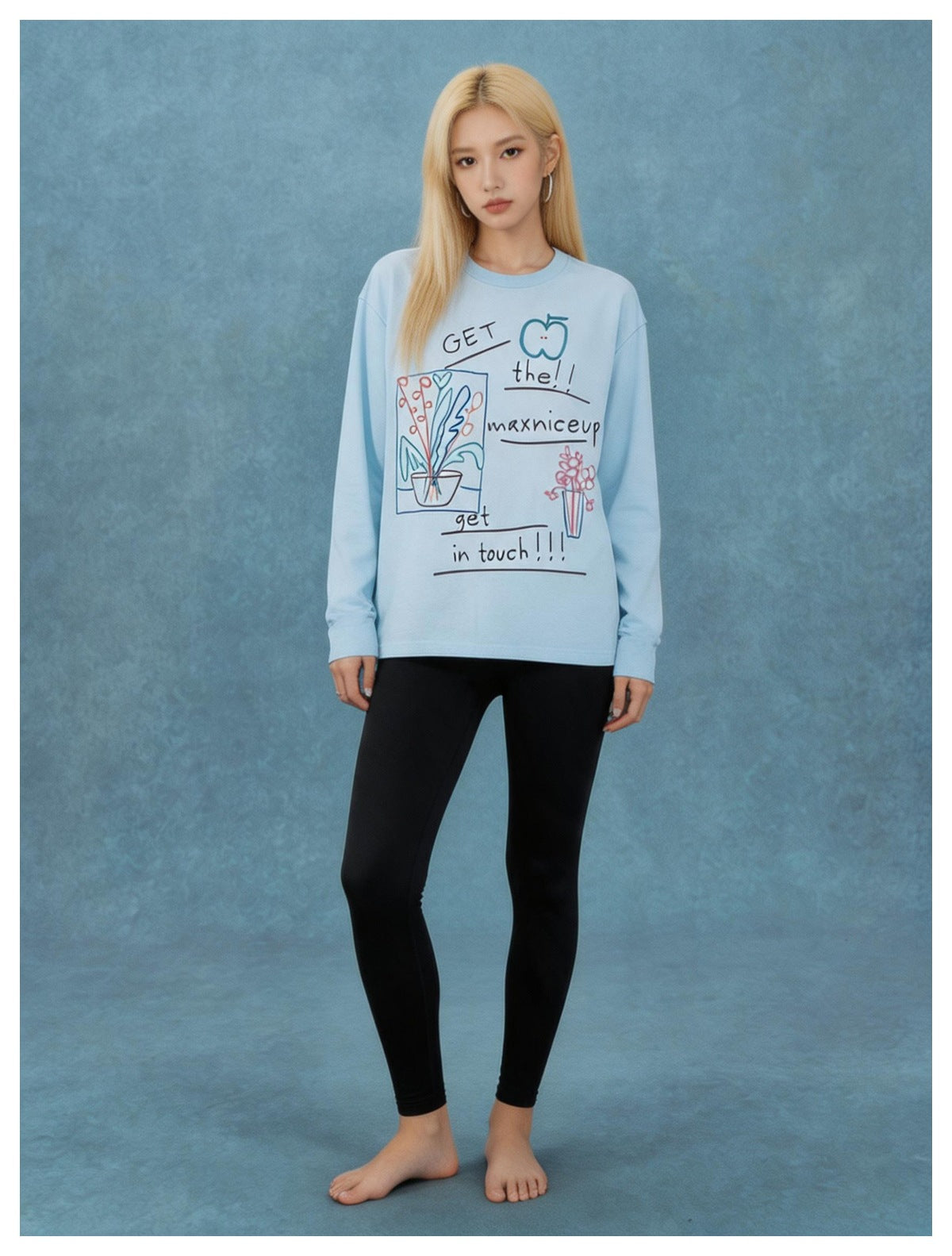 Model wearing light blue long-sleeve graphic tee with black leggings against a blue backdrop, full-length front view