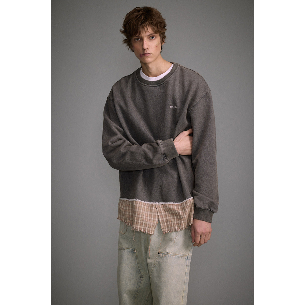 Man modeling a charcoal oversized crewneck sweatshirt with a frayed plaid lower panel over loose pale jeans.