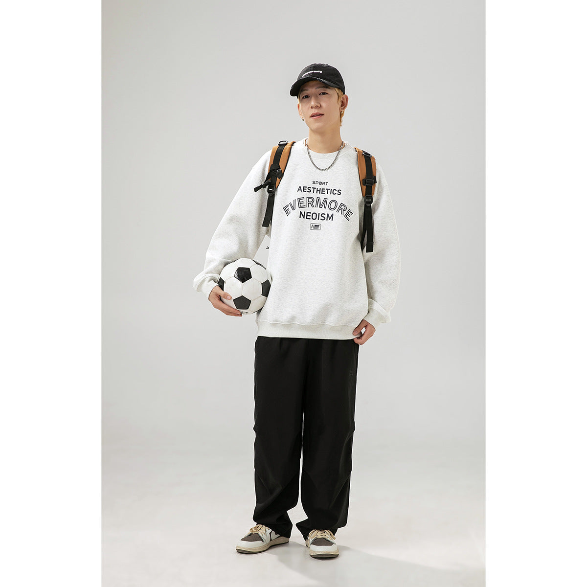 Model wearing a light gray crewneck sweatshirt with dark chest lettering, black pants, and a backpack, holding a soccer ball.