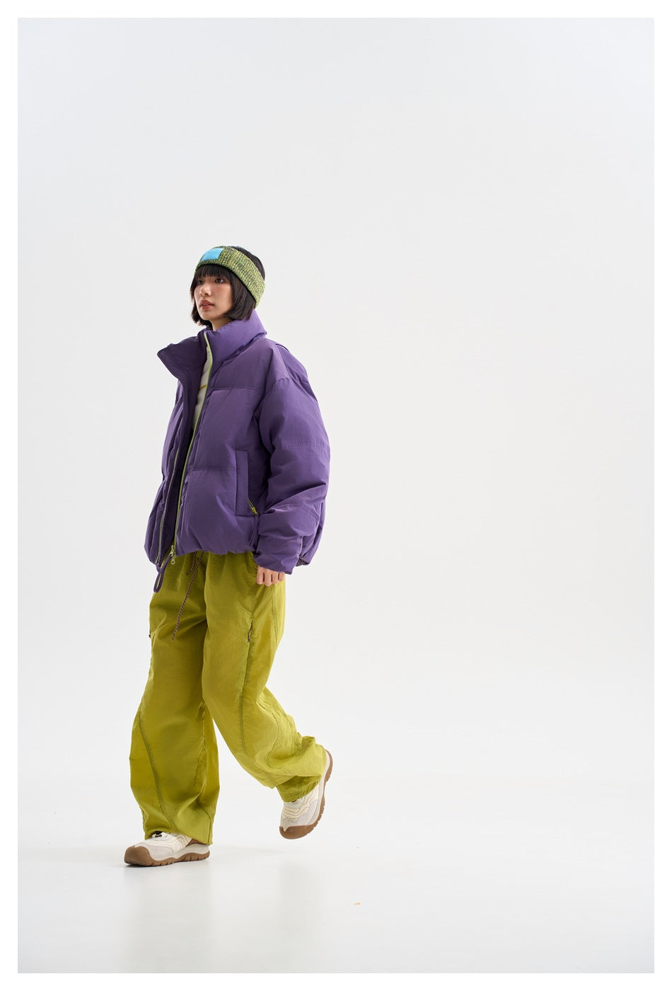 Side view of a model in a purple puffer jacket with lime zipper and loose lime trousers, walking in a studio.