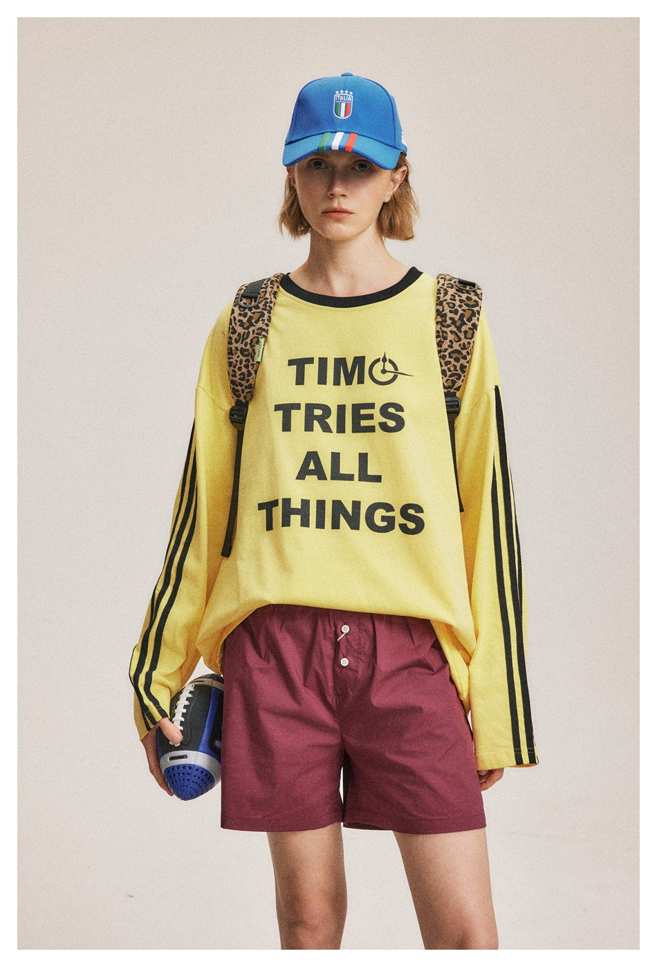 Front view of a model wearing a yellow shirt with black sleeve stripes and lettering, burgundy shorts, and a blue cap.