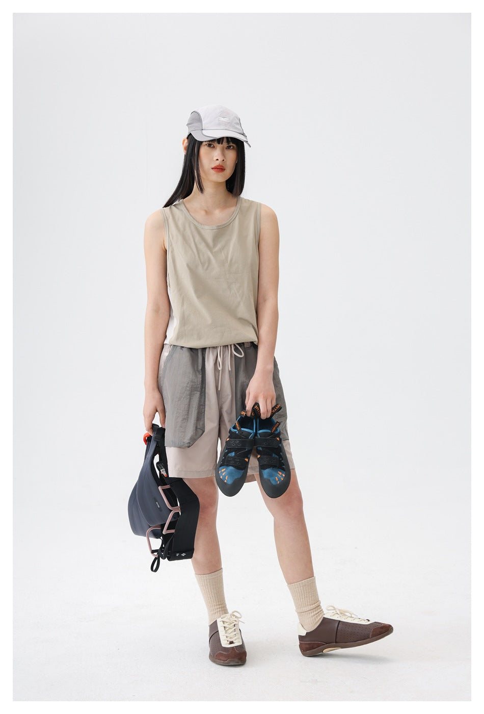 Model wearing a light beige sleeveless tank and two-tone gray drawstring shorts, holding outdoor gear.