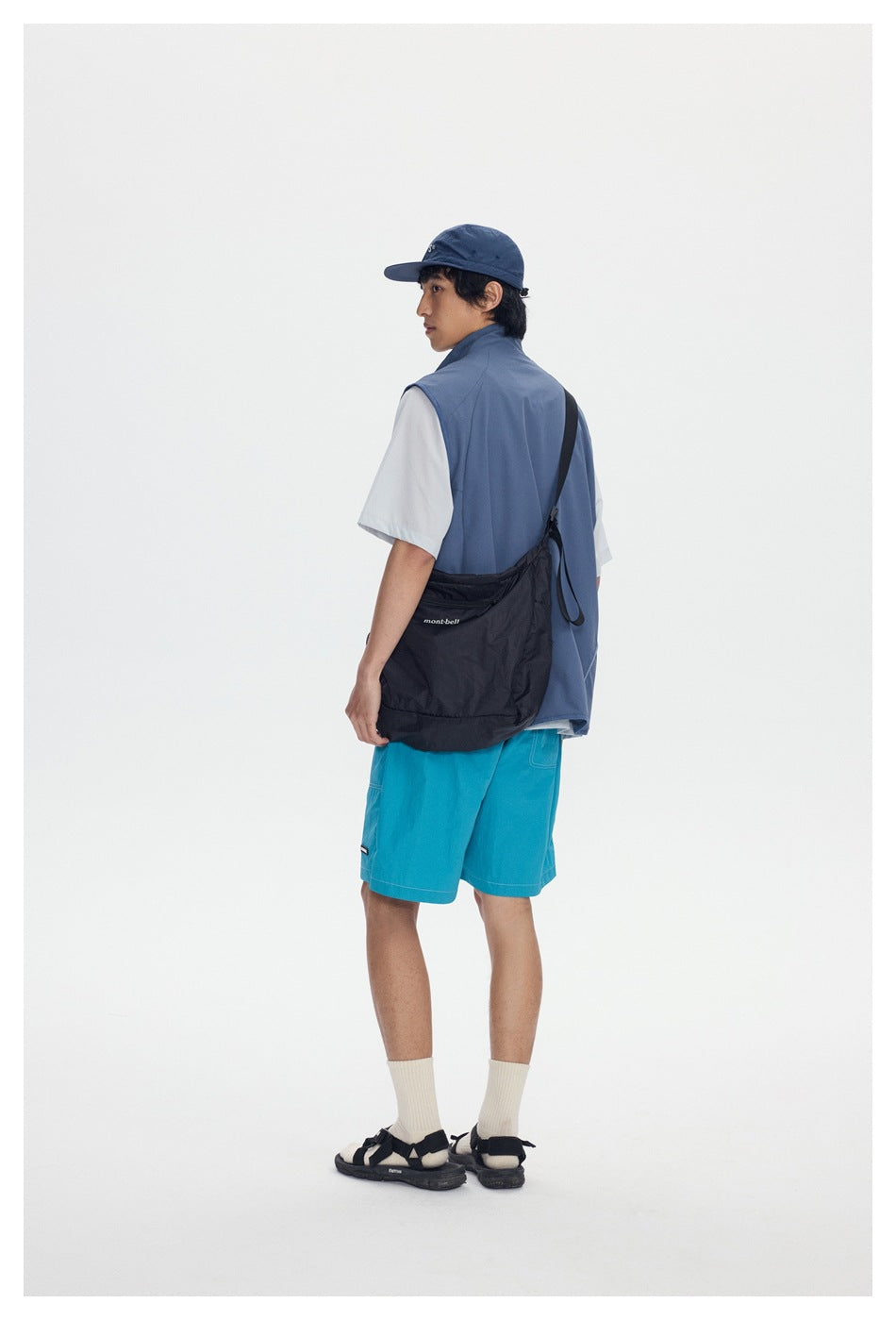 Rear view of a model in a slate-blue vest and turquoise shorts, carrying a black shoulder bag.
