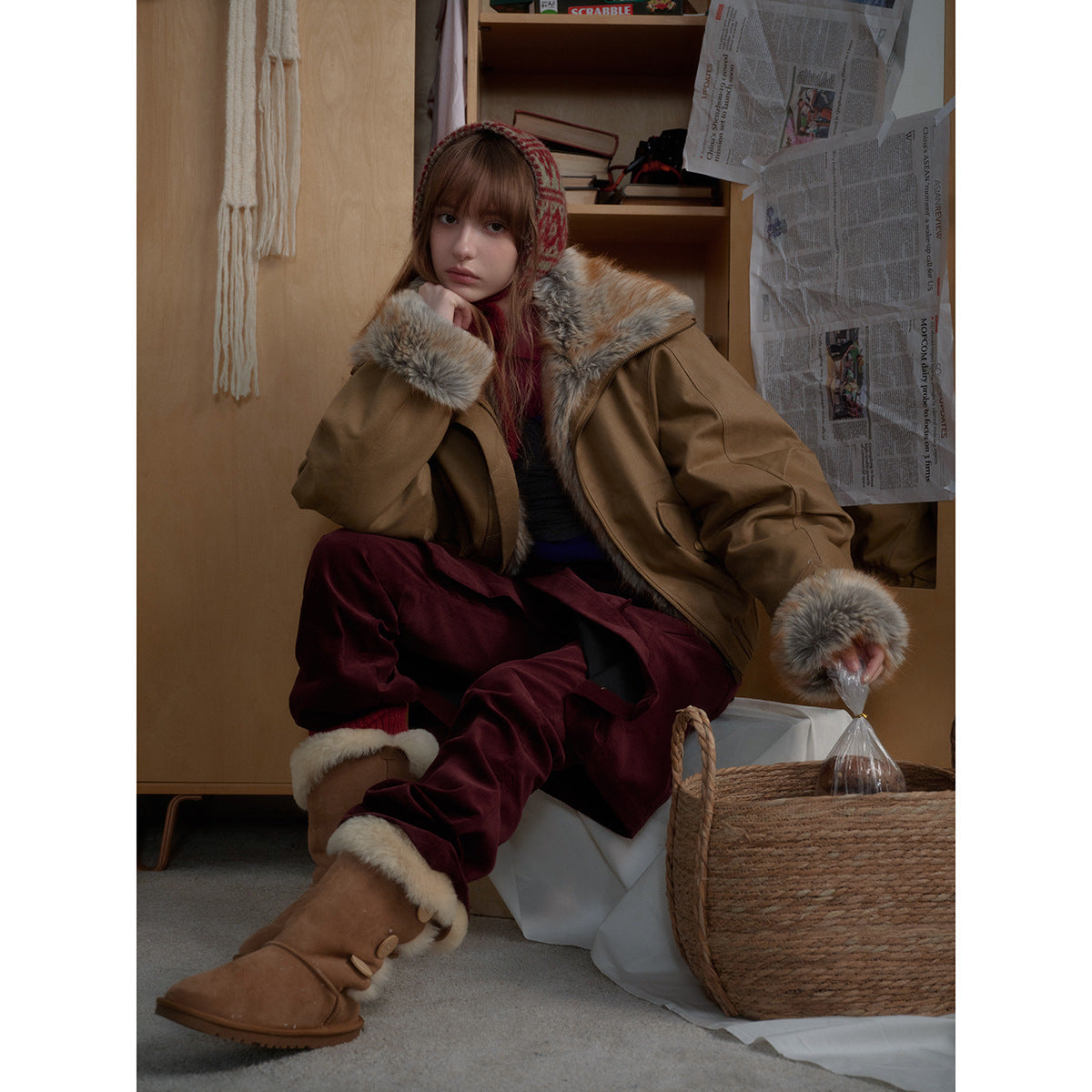 Seated model wearing an open tan jacket with plush fur trim at the collar and cuffs, paired with burgundy trousers.