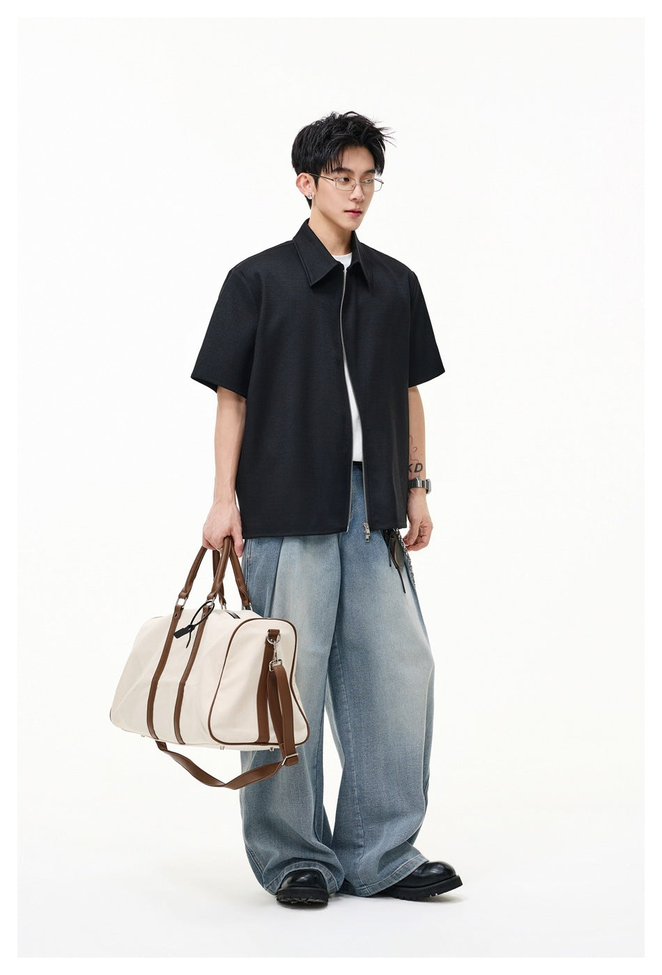 Full-length model in a black short-sleeve collared zip-front shirt and loose blue jeans, carrying a light-colored bag.