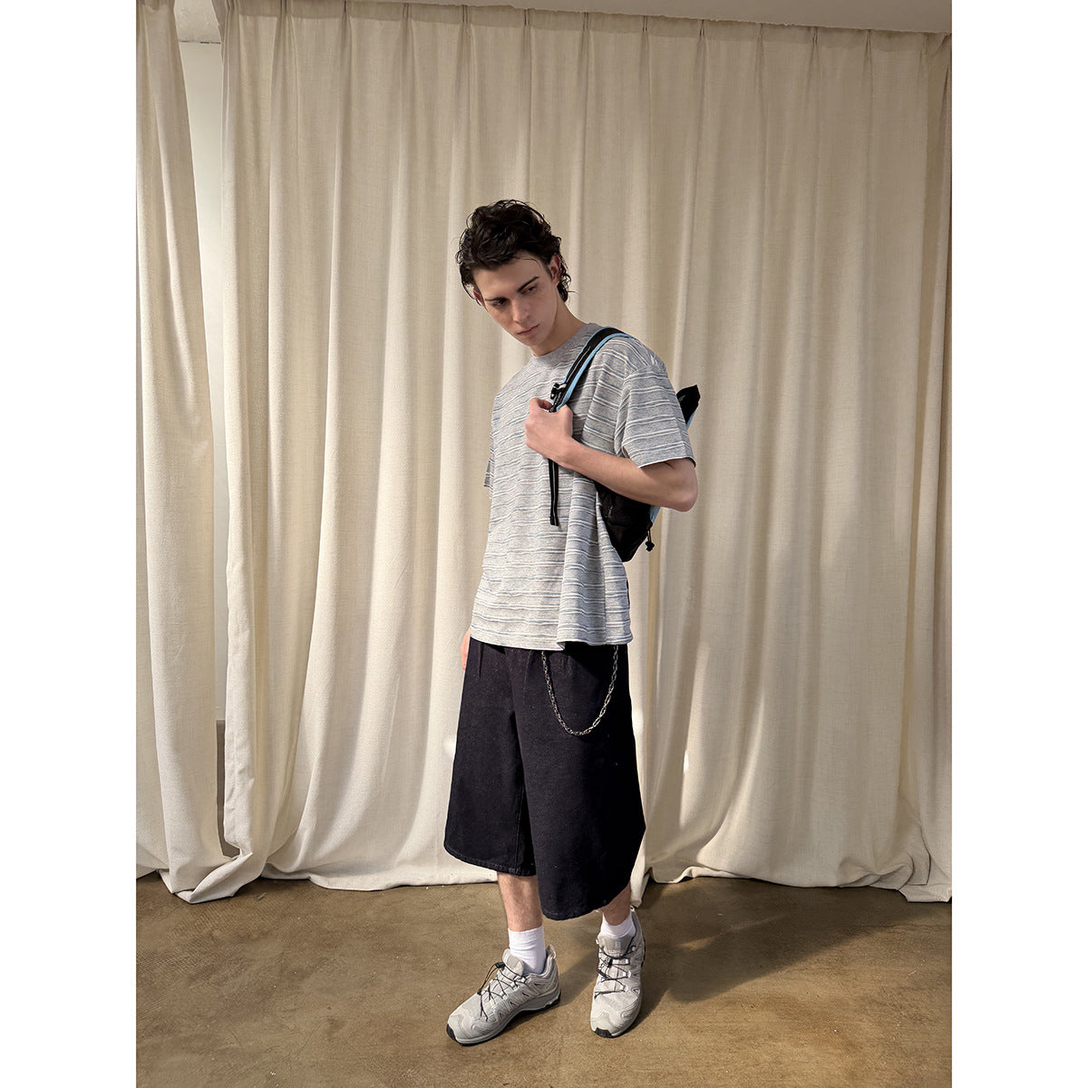 Model wearing a gray striped T-shirt and black knee-length shorts, shown at an angle with a shoulder bag.