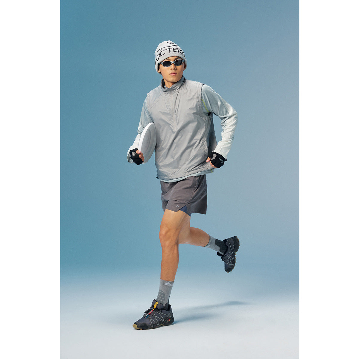 A man models a light gray pullover with long sleeves and dark running shorts, holding a flying disc.