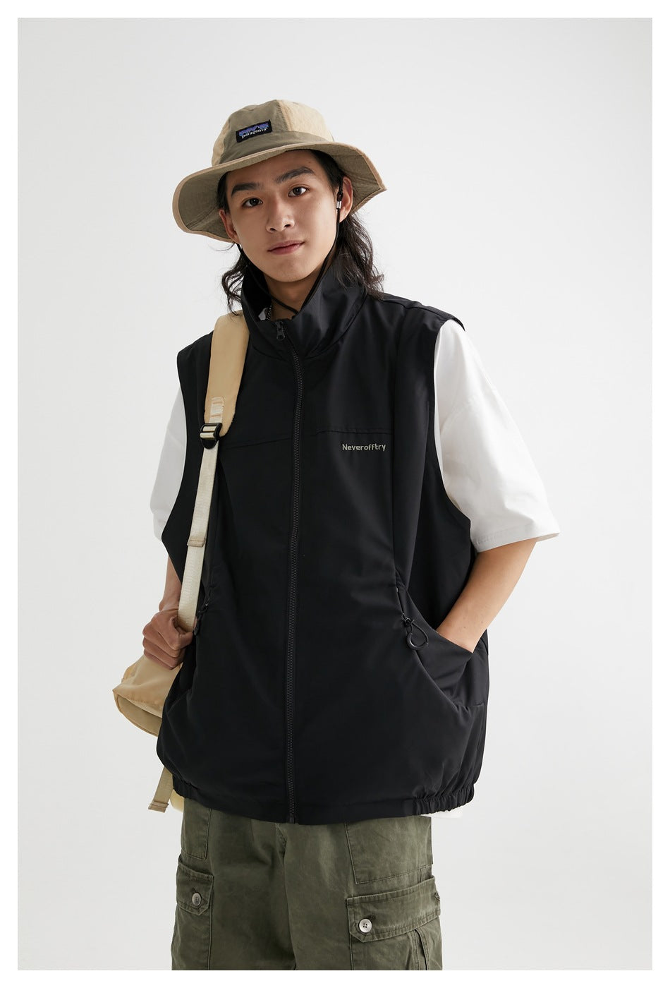 Model in a black zip-front vest with drawcord pockets, worn over a white T-shirt; a tan hat and backpack complete the outfit.